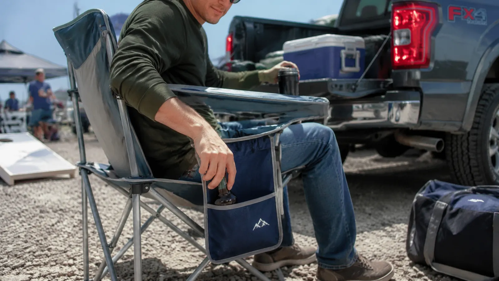 A person sitting in the camping chair at a tailgate, reaching into the side pouch for a drink while friends gather near a pickup truck.